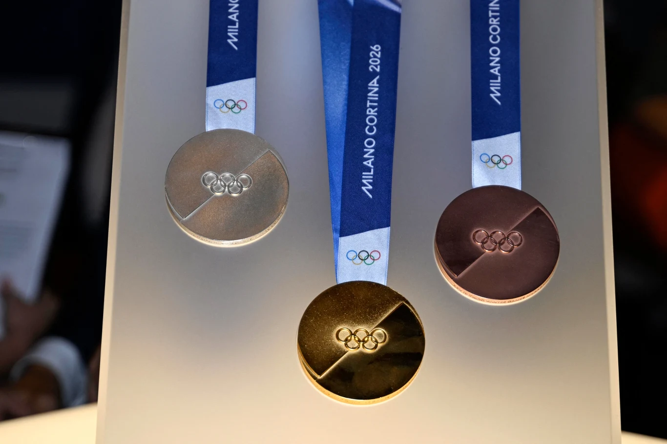 Trzy medale olimpijskie w barwach złota, srebra i brązu zawieszone na niebieskich wstążkach z napisem Milano Cortina 2026 oraz logo olimpijskie na każdym z nich.