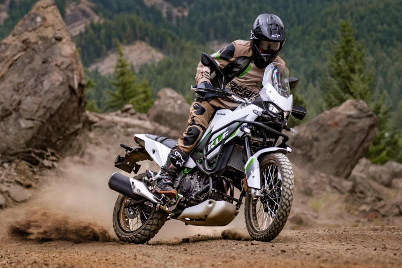 Po latach nieobecności Kawasaki wskrzesza model KLE. Motocyklista w pełnym stroju ochronnym jadący terenowym motocyklem po kamienistej, górskiej ścieżce. W tle widoczne są duże skały oraz zielone drzewa iglaste, tworzące surowy krajobraz.