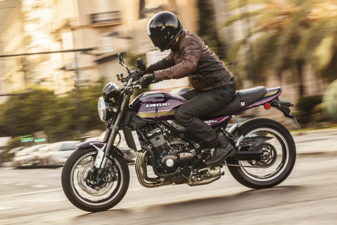 Klasyczna do bólu linia Z900RS nadal prezentuje się wyśmienicie. Motocyklista w czarnym kasku i brązowej kurtce jedzie klasycznym motocyklem Kawasaki w ruchu miejskim. Dynamiczne ujęcie z rozmytym tłem podkreślającym prędkość pojazdu.