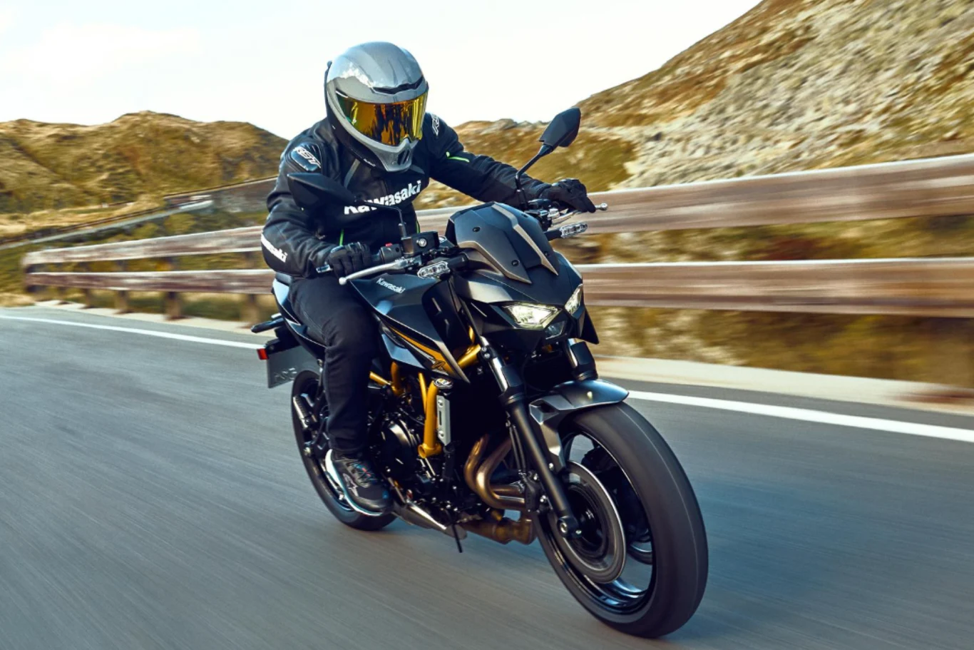 Naked ma być jeszcze bardziej komfortowy, ale przy tym wyjątkowo drapieżny. Motocyklista w nowoczesnym kasku i kombinezonie na czarnym motocyklu sportowym pokonuje krętą górską drogę z barierkami ochronnymi, w tle nachylone zbocza i skały.