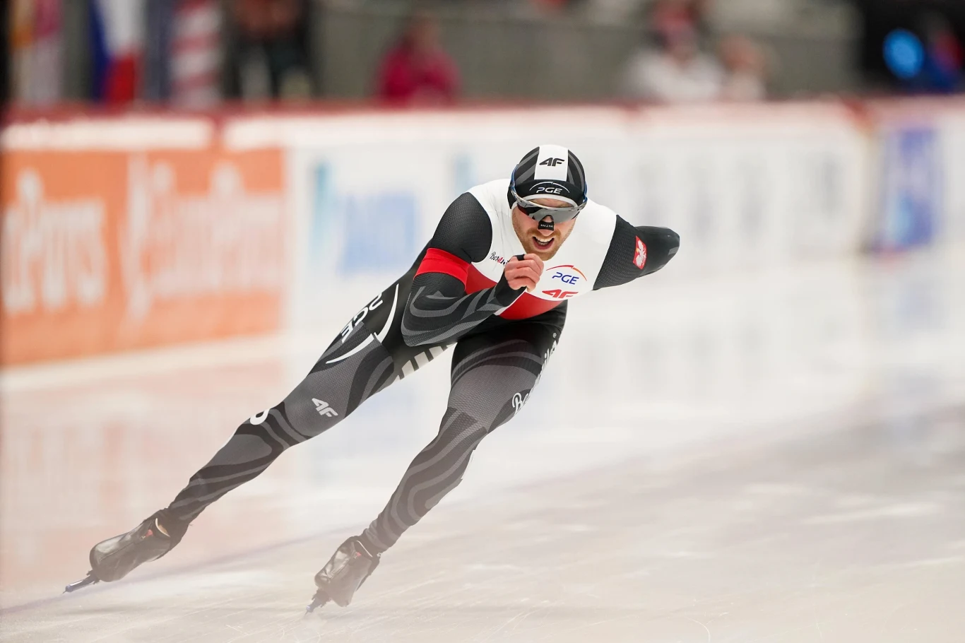 Damian Żurek jest wielką nadzieją Polski na medal zimowych igrzysk olimpijskich 2026 Łyżwiarz szybki w specjalnym stroju startuje na lodowisku podczas zawodów, dynamicznie pochylony do przodu, w pełnym skupieniu, wokół rozmyte tło areny sportowej.