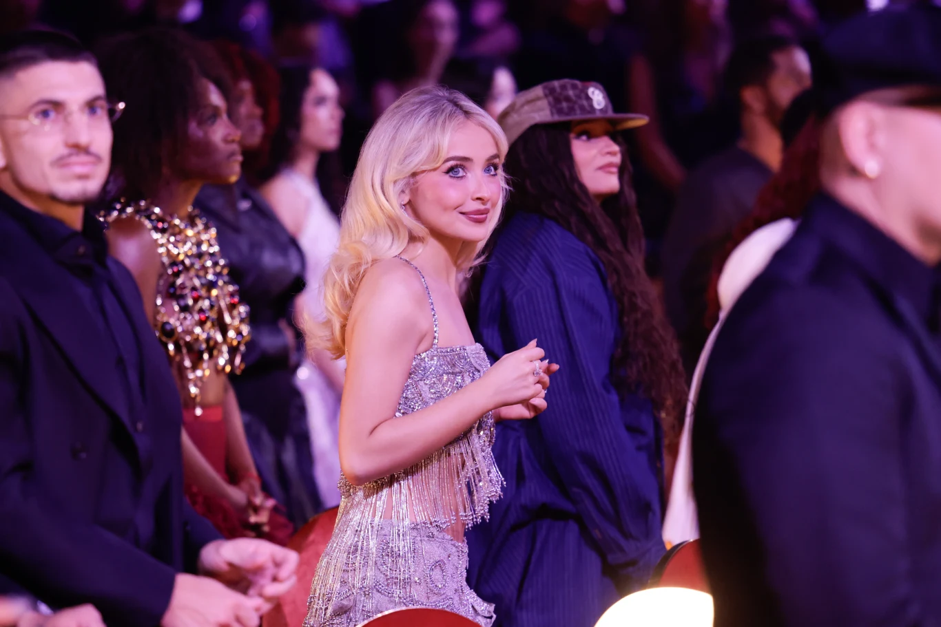 Sabrina Carpenter podczas gali rozdania nagród Grammy 2026 Kobieta z blond włosami w błyszczącej sukni stoi wśród grupy osób na eleganckiej gali, w tle widać innych gości w zróżnicowanych stylizacjach, wokół panuje podniosła i uroczysta atmosfera.