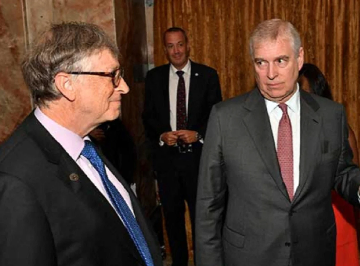 Dwóch mężczyzn w garniturach rozmawia ze sobą podczas oficjalnego wydarzenia w eleganckim wnętrzu, za nimi stoi mężczyzna w marynarce oraz zasłony i marmurowa ściana.