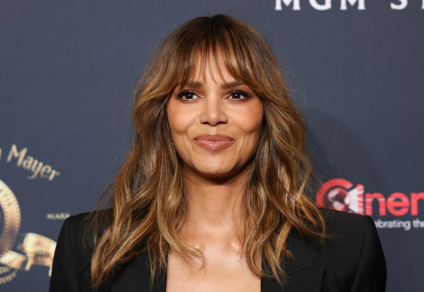 Halle Berry Kobieta z rozpuszczonymi, falującymi włosami o jasnych refleksach, ubrana w czarną marynarkę. W tle widoczne jest ciemne tło z logotypami i napisami, sugerujące wydarzenie branżowe lub galę.