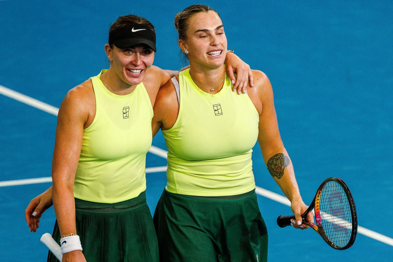 Dwie kobiety, ubrane w żółte koszulki sportowe i zielone spódnice, stoją obok siebie na korcie tenisowym. Jedna z nich trzyma rakietę tenisową, obie uśmiechnięte i obejmujące się ramionami, wyrażając radość po meczu.