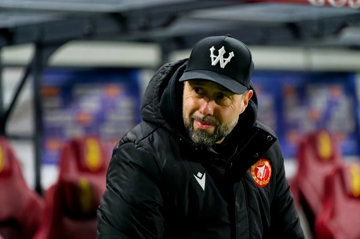 Mężczyzna w czarnej kurtce sportowej i czapce z daszkiem z literą W stoi przed ławką rezerwowych na tle stadionu. Na jego kurtce znajduje się czerwono-żółta naszywka, w tle zamazane miejsca siedzące.