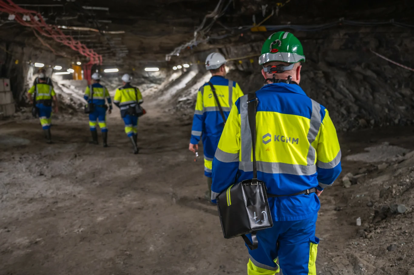 Górnicy z KGHM chcą podwyżek stawek płac zasadniczych Grupa pracowników ubranych w odblaskowe kombinezony i kaski ochronne, przemieszczających się korytarzem podziemnej kopalni, otoczenie stanowią skalne ściany oraz elementy infrastruktury górniczej.