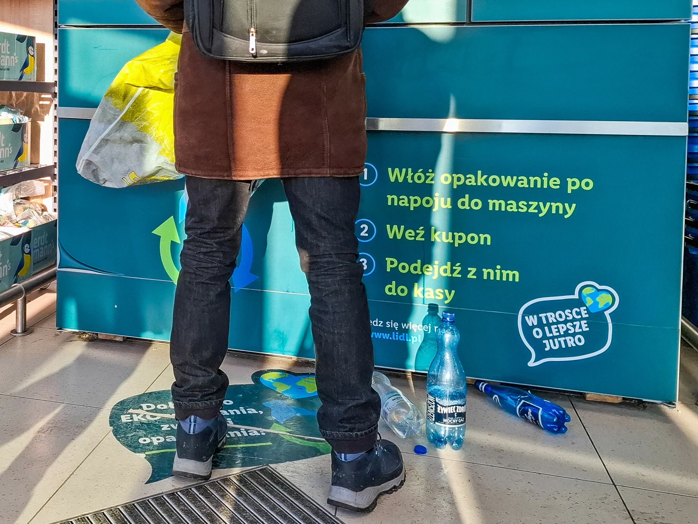 Na systemie kaucyjnym niektórzy zarabiają po kilkaset złotych tygodniowo Mężczyzna stojący przed maszyną do zwrotu butelek, obok na podłodze plastikowe butelki, na tablicy instrukcje dotyczące procesu zwrotu oraz ekologia.