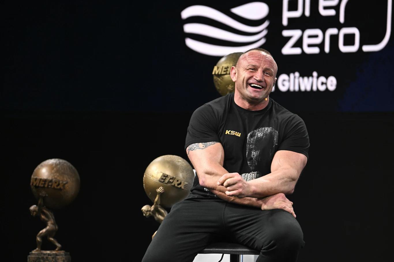 Mężczyzna o umięśnionej sylwetce siedzi na krześle z szerokim uśmiechem na twarzy, na tle czarnym z kulami MET-RX oraz napisem Gliwice i logo firmy w tle.