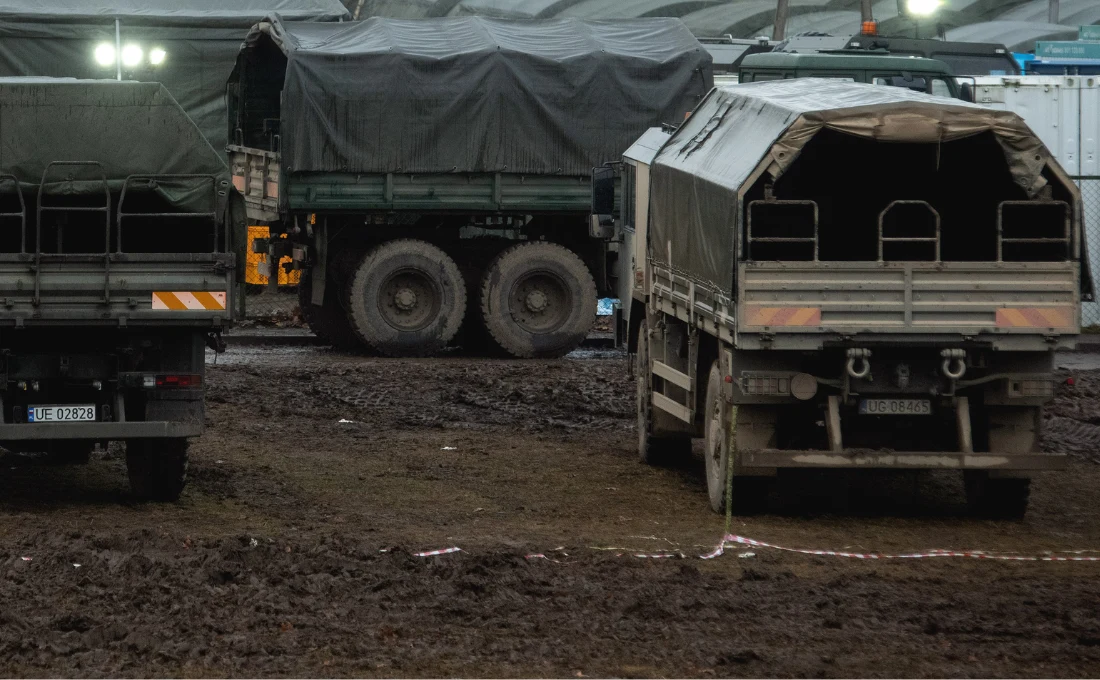 Kilka wojskowych ciężarówek z plandekami, ustawionych na błotnistym terenie, w otoczeniu prowizorycznych świateł i zabudowań, bez widocznych ludzi.