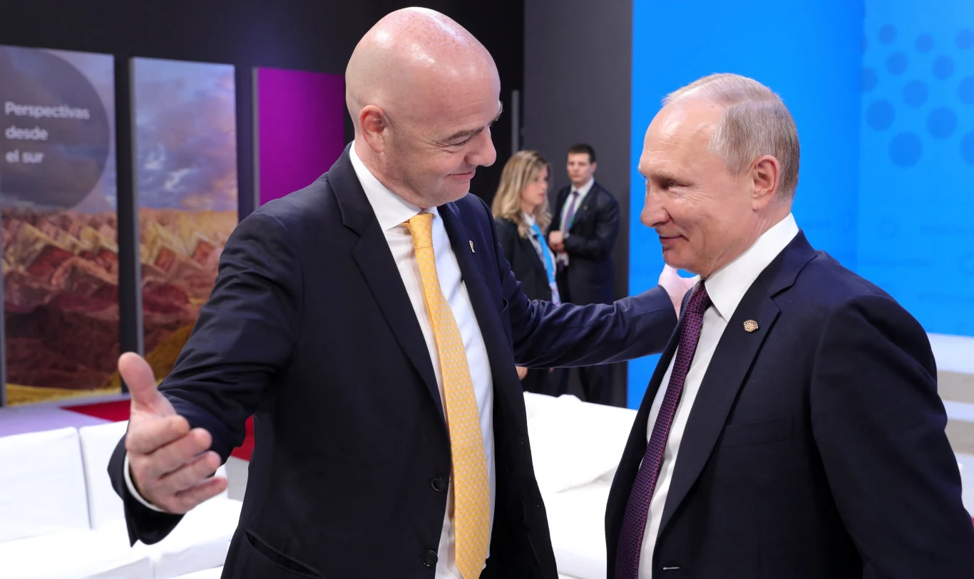 Dwóch mężczyzn w garniturach stoi naprzeciw siebie w przyjaznej atmosferze, jeden z nich z rozłożonymi rękami jakby zapraszał do uścisku, w tle kilka osób i kolorowe panele wystawowe.