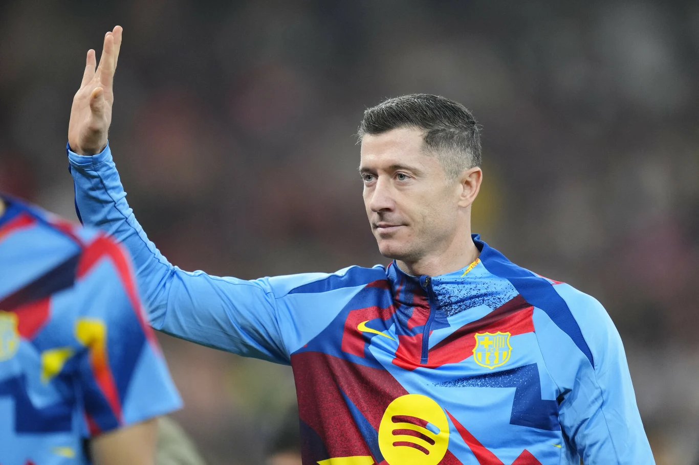 Piłkarz w niebieskiej koszulce z logotypem FC Barcelona oraz sponsora, unosi rękę w geście powitania lub pozdrowienia. W tle widoczna sylwetka innego zawodnika oraz rozmazane trybuny stadionu.