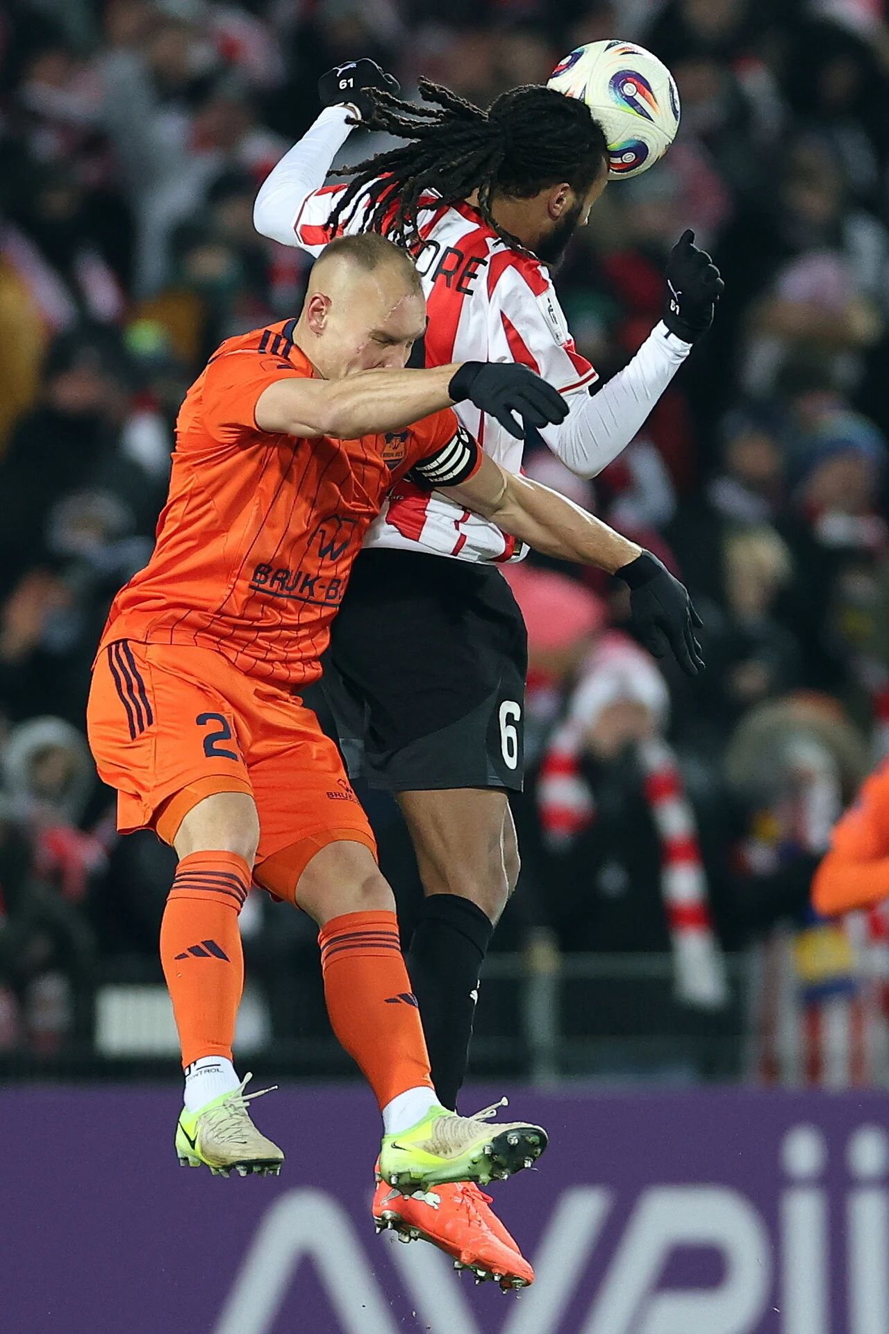 Dwóch piłkarzy walczy o piłkę w powietrzu podczas meczu piłkarskiego, jeden z zawodników ubrany jest w pomarańczowy strój, drugi w biało-czerwone paski; wokół nich widać zamazany tłum kibiców.