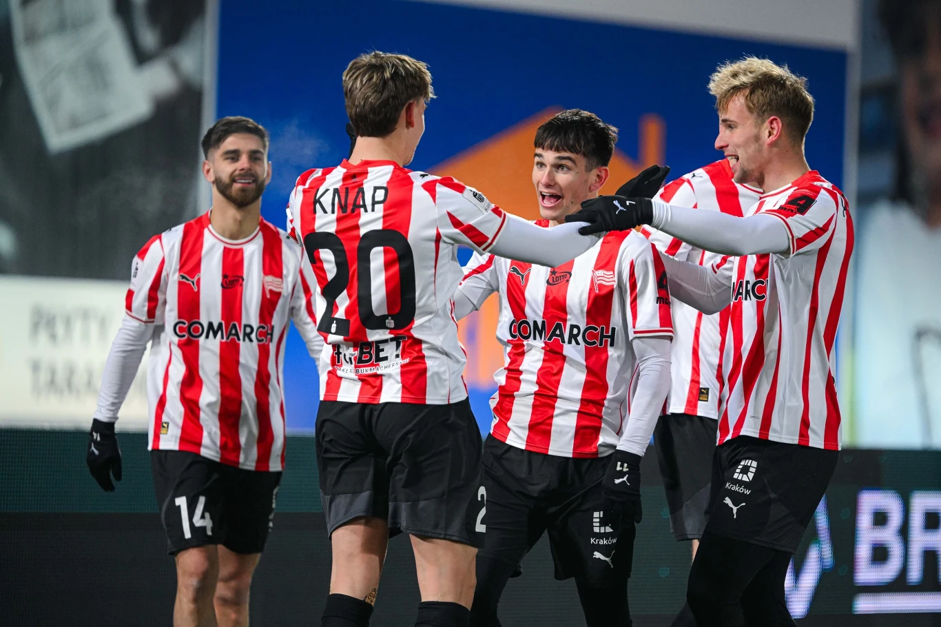 Grupa piłkarzy w biało-czerwonych pasach świętuje zdobycie gola na boisku, kilku z nich obejmuje się nawzajem z widoczną radością na twarzach. W tle niewyraźne napisy oraz fragment trybun stadionu.