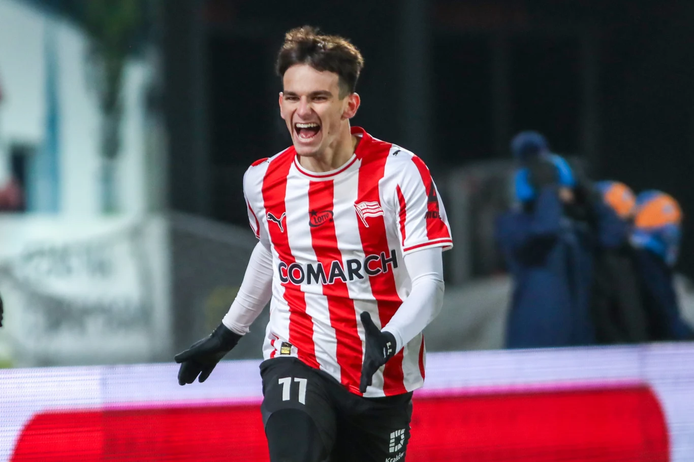 Piłkarz w biało-czerwonej pasiasto-czerwonej koszulce z radością biegnie po murawie, wykonując gest triumfu, w tle rozmazane postaci i elementy stadionu.