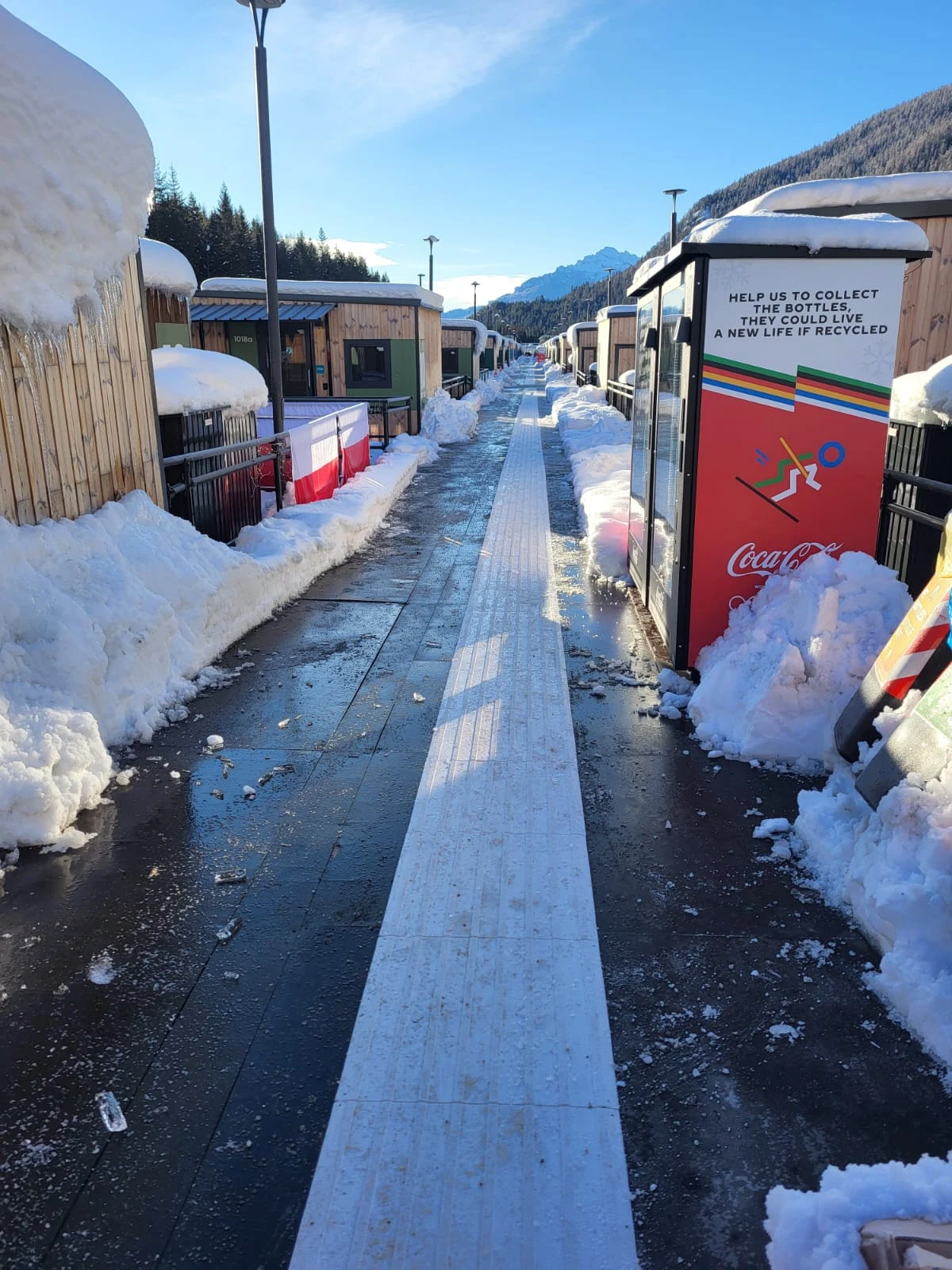 Pokryta śniegiem alejka z wyraźnie odśnieżonym chodnikiem prowadzi przez strefę z zadaszonymi budkami. Z obu stron ścieżki wysokie zaspy śniegu oraz czerwony automat do napojów Coca-Cola po prawej stronie. W tle widoczne są góry oraz niebo, a całość oś...