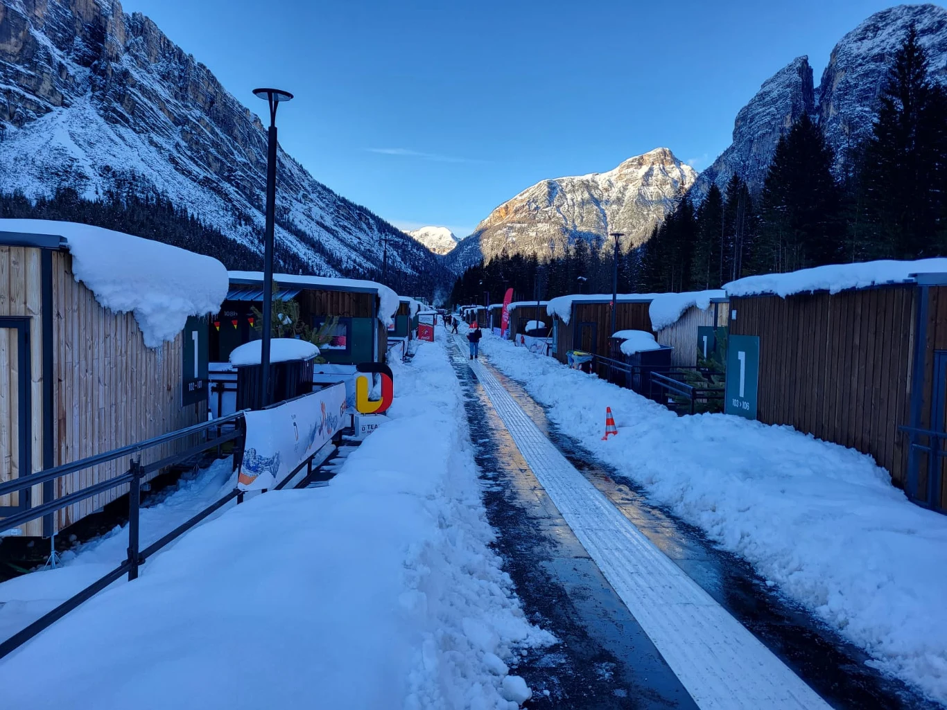 Zaśnieżona ścieżka między drewnianymi budynkami, z wysokimi górami w tle i błękitnym niebem. Po obu stronach widać śnieżne daszki i zalesione stoki, na końcu drogi wyłania się łagodne światło słoneczne oświetlające szczyty.