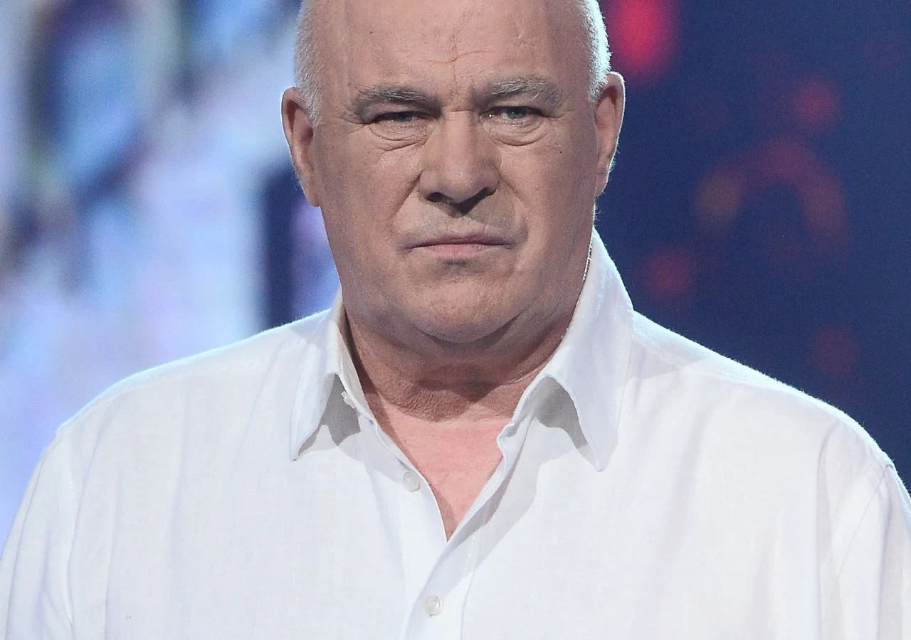Ryszard Rynkowski Starszy mężczyzna o poważnym wyrazie twarzy, ubrany w białą koszulę, stoi na rozmytym tle z elementami kolorowych świateł.