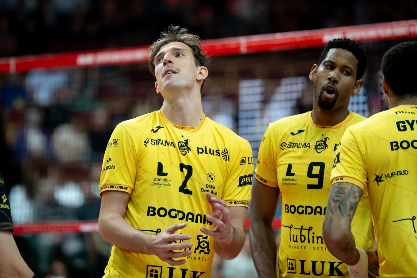 Grupa siatkarzy w żółtych koszulkach z nazwiskami oraz numerami, stojących przy siatce w hali sportowej. Dwóch z nich znajduje się na pierwszym planie, skupiają wzrok i uwagę, trzymając ręce w widocznym geście.