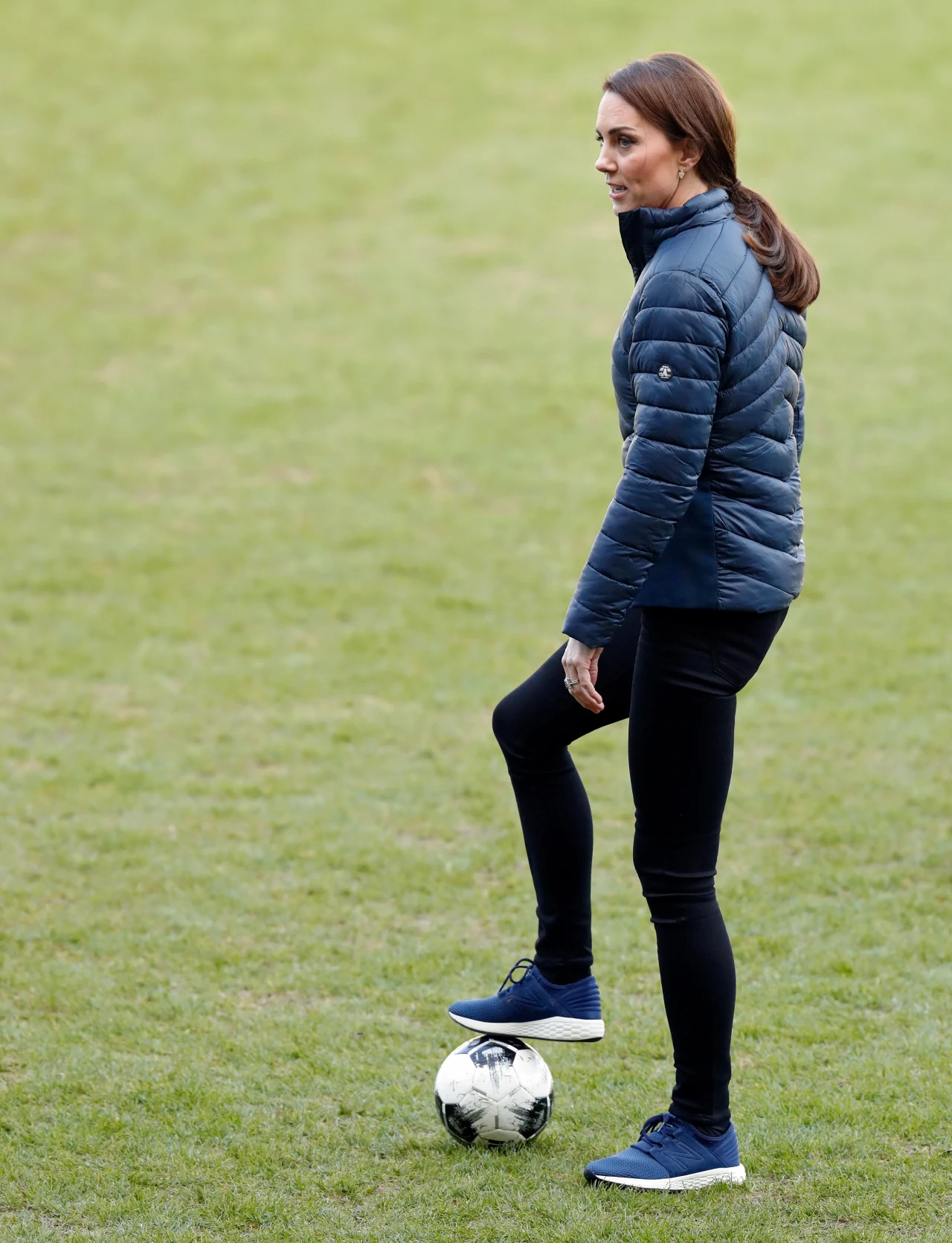 Kobieta ubrana w sportową kurtkę, czarne spodnie i ciemne buty stoi na zielonej murawie z jedną nogą opartą na piłce nożnej.