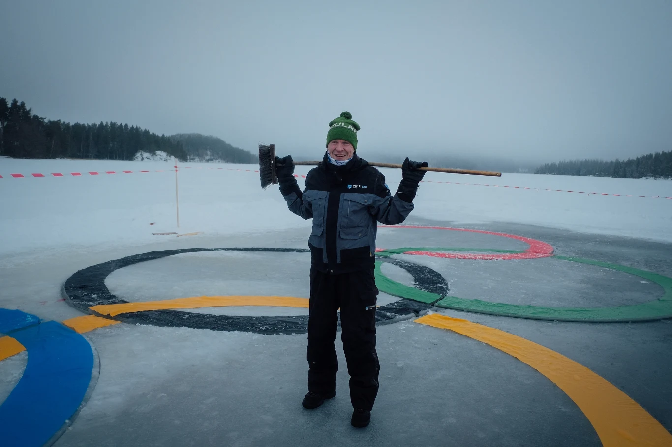 Uśmiechnięta osoba w zimowym ubraniu stoi na zamarzniętym jeziorze, na lodzie widoczne są namalowane kolorowe olimpijskie pierścienie, w tle ośnieżone drzewa i zamglony krajobraz. Osoba trzyma dużą szczotkę na ramionach.