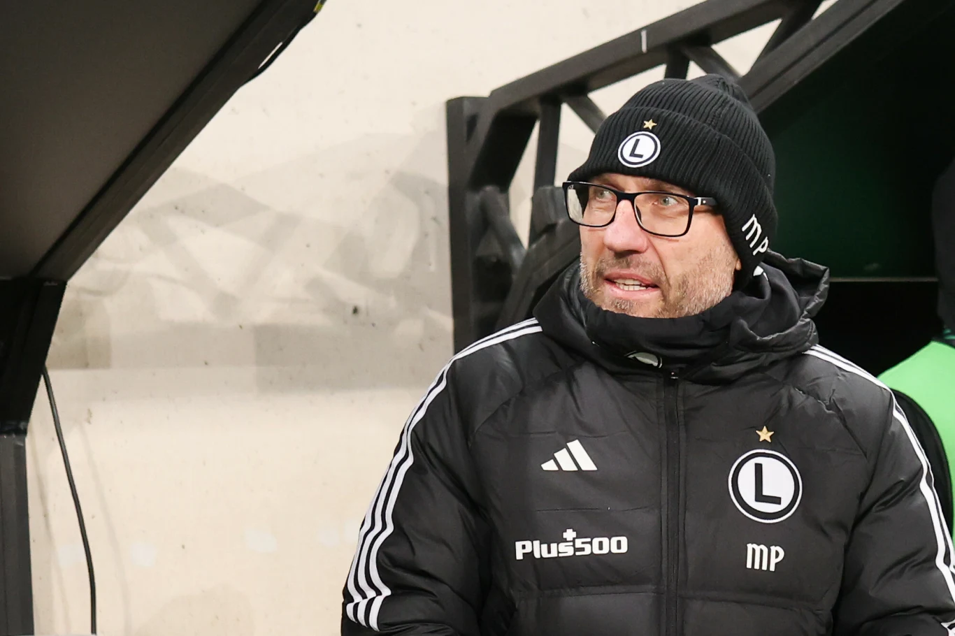 Marek Papszun Mężczyzna w sportowej kurtce i czapce z logo Legii Warszawa, ubrany zimowo, stoi obok zadaszenia stadionowego, w tle jasna ściana.