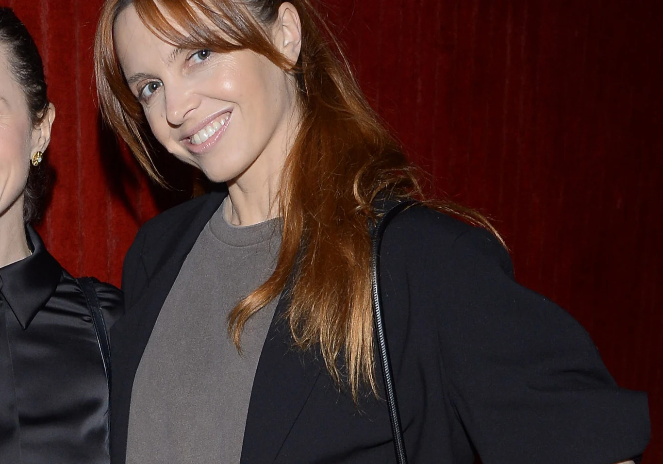 Uśmiechnięta kobieta z rudymi włosami ubrana w szarą bluzkę i czarną marynarkę na tle czerwonej zasłony.