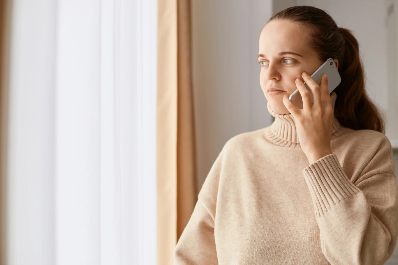Kobieta w jasnym swetrze rozmawia przez telefon, stoi przy oknie z zasłoną i patrzy zamyślona w dal.