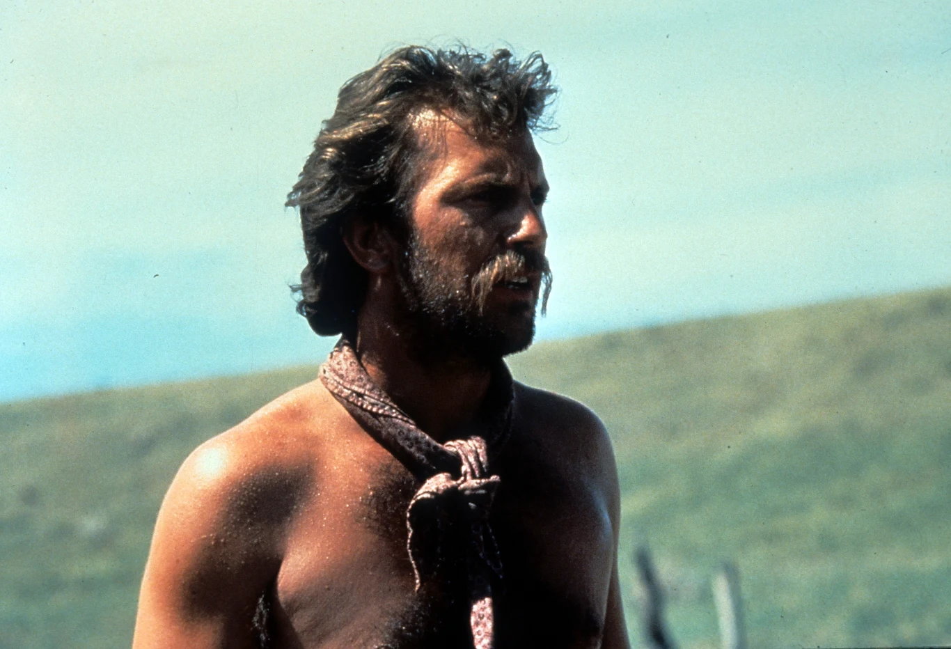 Mężczyzna z brodą i rozwichrzonymi włosami, z odsłoniętymi ramionami oraz sznurem zawiązanym wokół szyi, stoi na tle rozmytego krajobrazu z zielonymi wzgórzami.