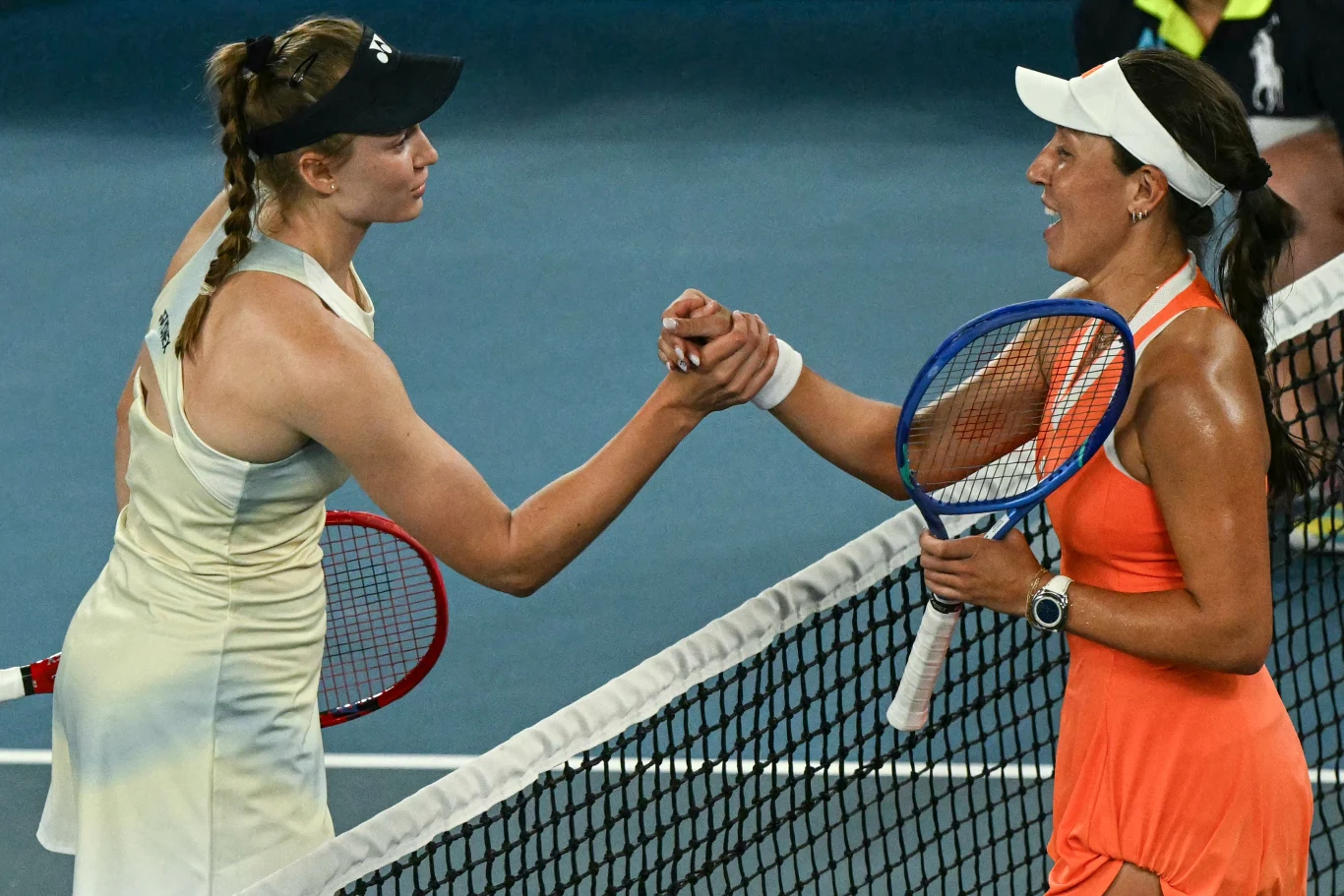 Dwie tenisistki ściskają sobie dłonie nad siatką na kortach tenisowych po zakończeniu meczu, obie trzymają rakiety tenisowe, jedna z zawodniczek ubrana jest na biało, druga w pomarańczową sukienkę sportową, atmosfera jest fair play.