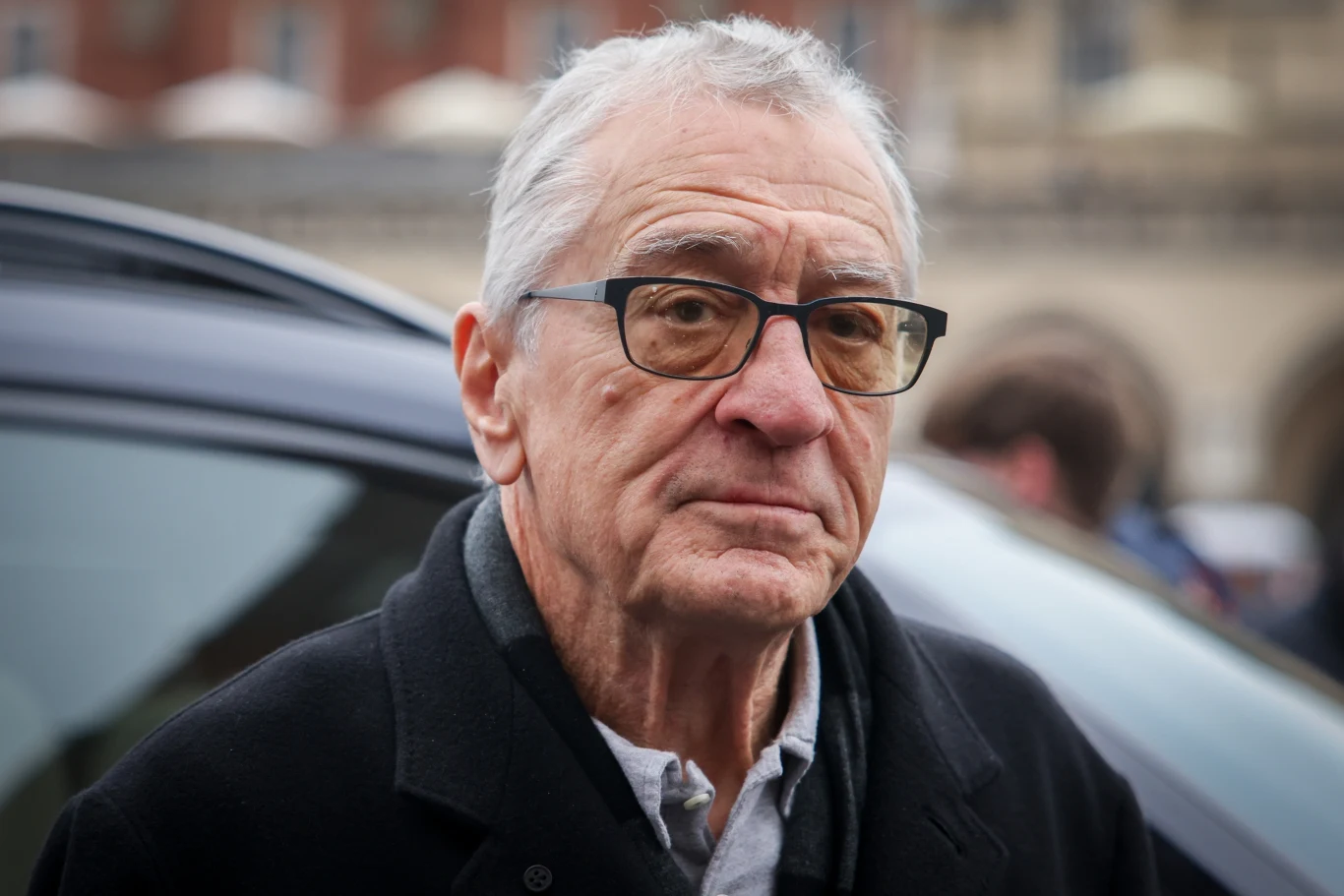 Starszy mężczyzna w okularach i ciemnym płaszczu stoi na zewnątrz, w tle widoczne nieostre budynki i samochód.