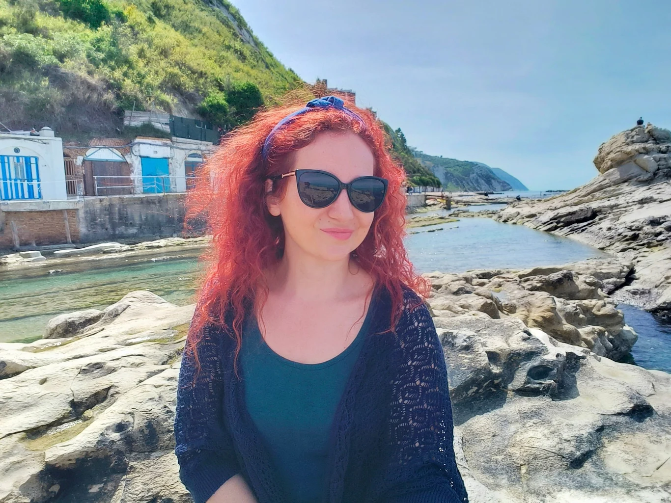 Kobieta z rudymi, kręconymi włosami i ciemnymi okularami przeciwsłonecznymi siedzi na skalistym wybrzeżu. W tle akwatyczny krajobraz, wzgórza porośnięte roślinnością oraz budynki z niebiesko-białymi drzwiami.