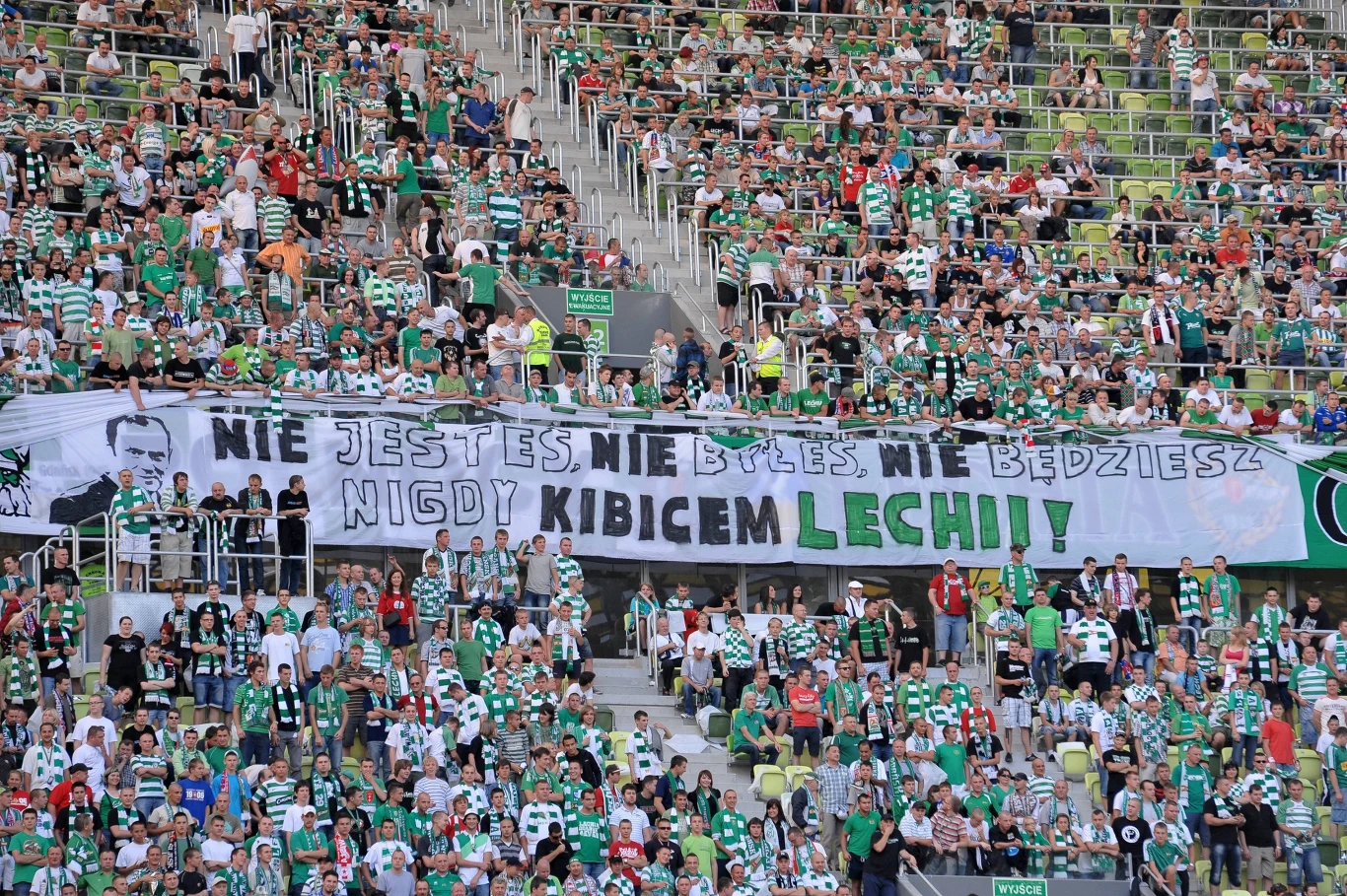 Tłum kibiców ubranych w biało-zielone barwy zajmuje trybuny stadionu, trzymając duży transparent z napisem 'Nie jesteś, nie byłeś, nie będziesz nigdy kibicem Lechii'. Atmosfera entuzjastyczna, wiele osób śpiewa i macha szalikami.
