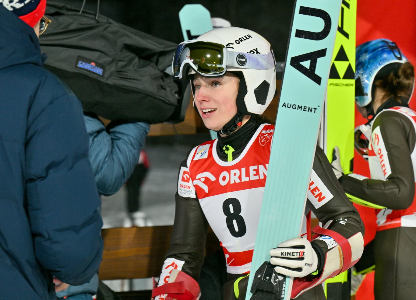 Sportsmenka w kasku narciarskim i goglach trzymająca narty, ubrana w kombinezon startowy z numerem 8. W tle widać innych sportowców oraz elementy infrastruktury zawodów narciarskich.