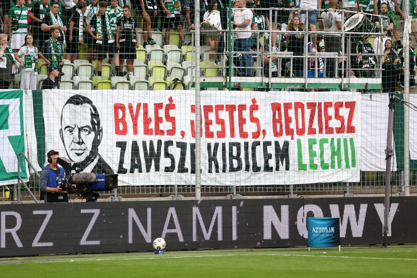 Tłum kibiców na stadionie piłkarskim, duży baner z hasłem kibicowskim i podobizną mężczyzny, dominują barwy zielone i białe, część ludzi ubrana w klubowe stroje, przed ogrodzeniem ustawiona jest kamera telewizyjna i piłka.