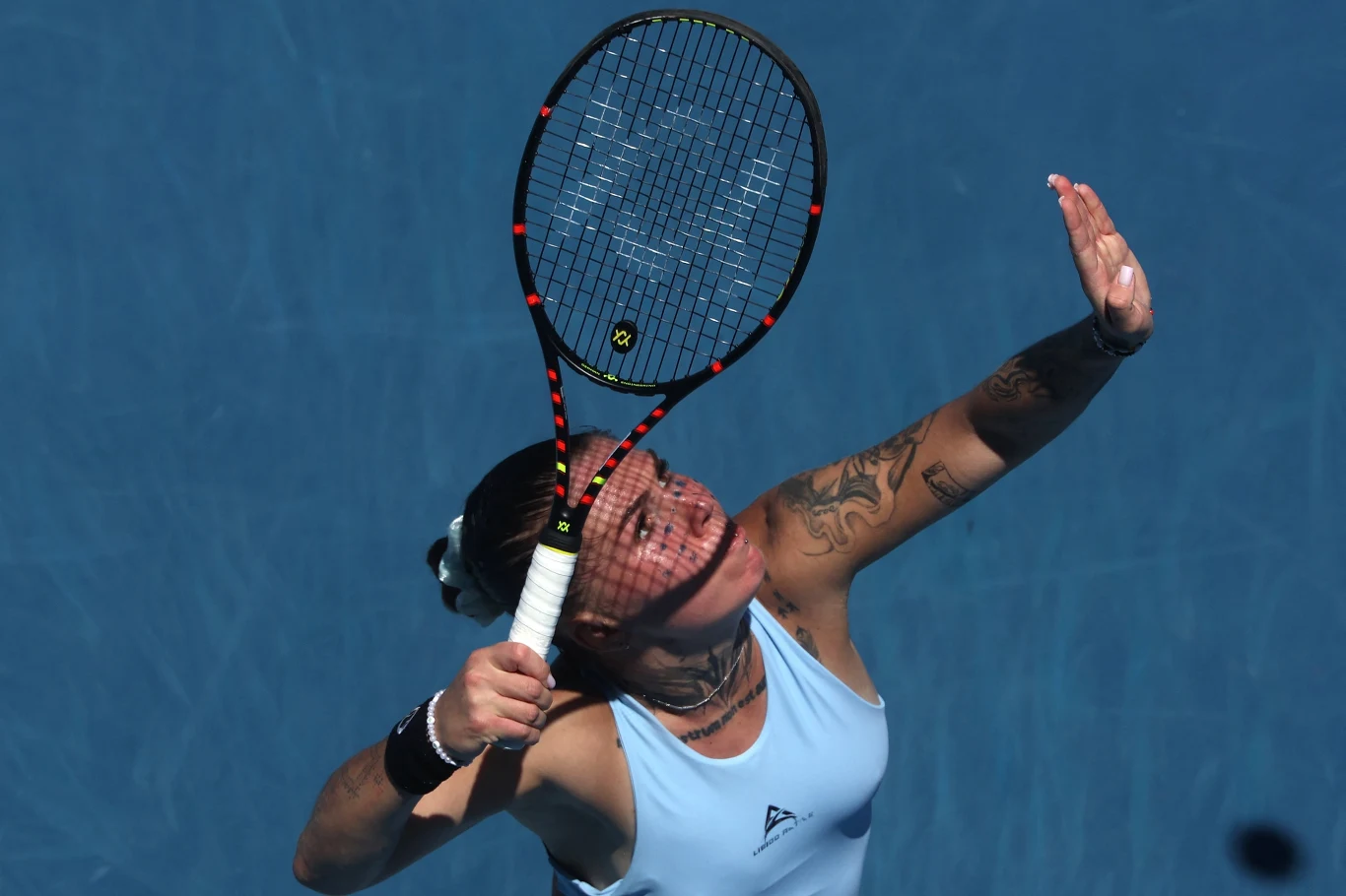 Tenisistka z tatuażami wykonuje serwis podczas meczu na kortach z niebieską nawierzchnią. Dynamiczny ruch rakietą, skupienie na twarzy zawodniczki, jasny sportowy strój.