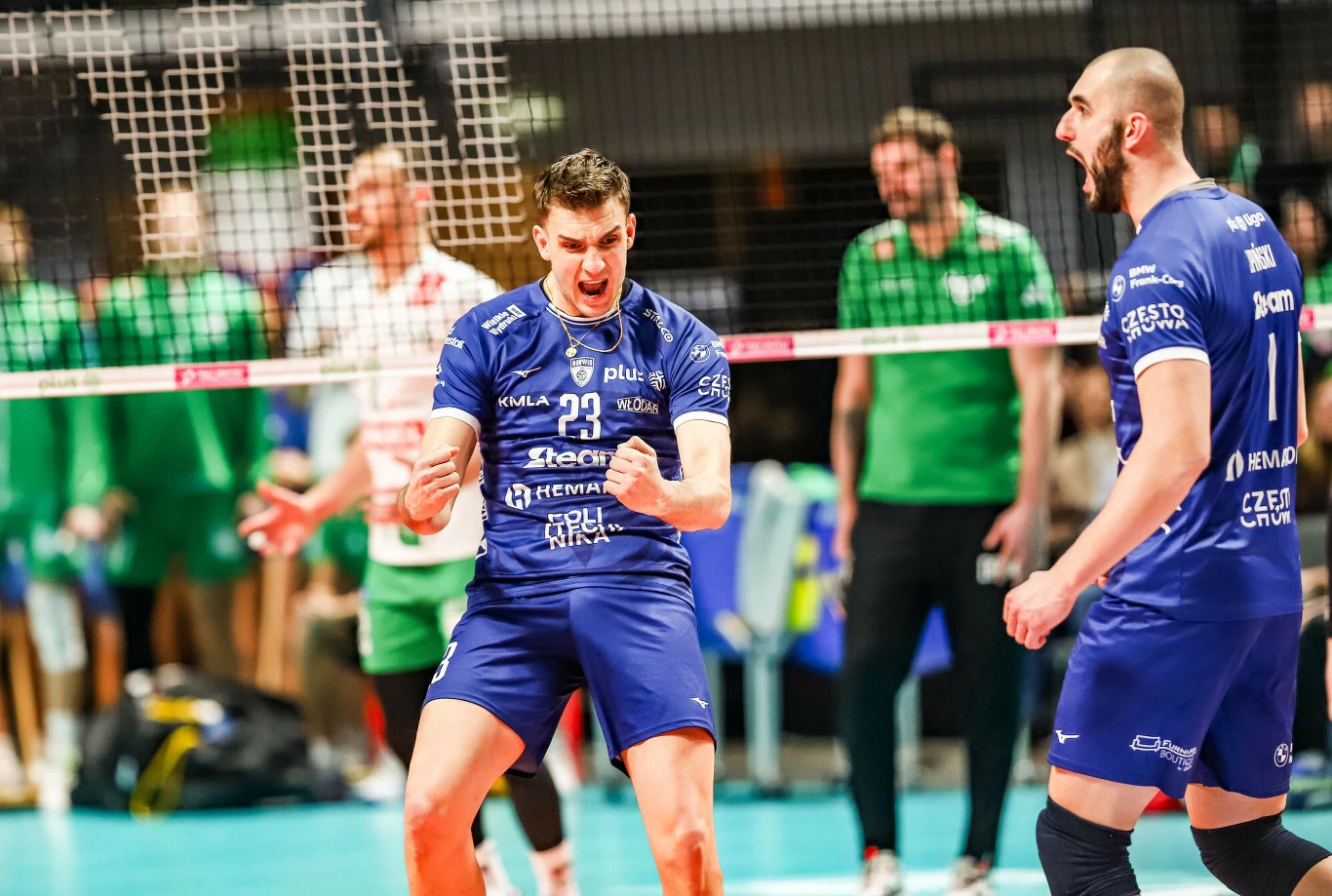 Dwóch siatkarzy w niebieskich strojach wyraża silne emocje radości i triumfu po udanej akcji na boisku, w tle kilku zawodników drużyny przeciwnej oraz siatka oddzielająca obie strony.