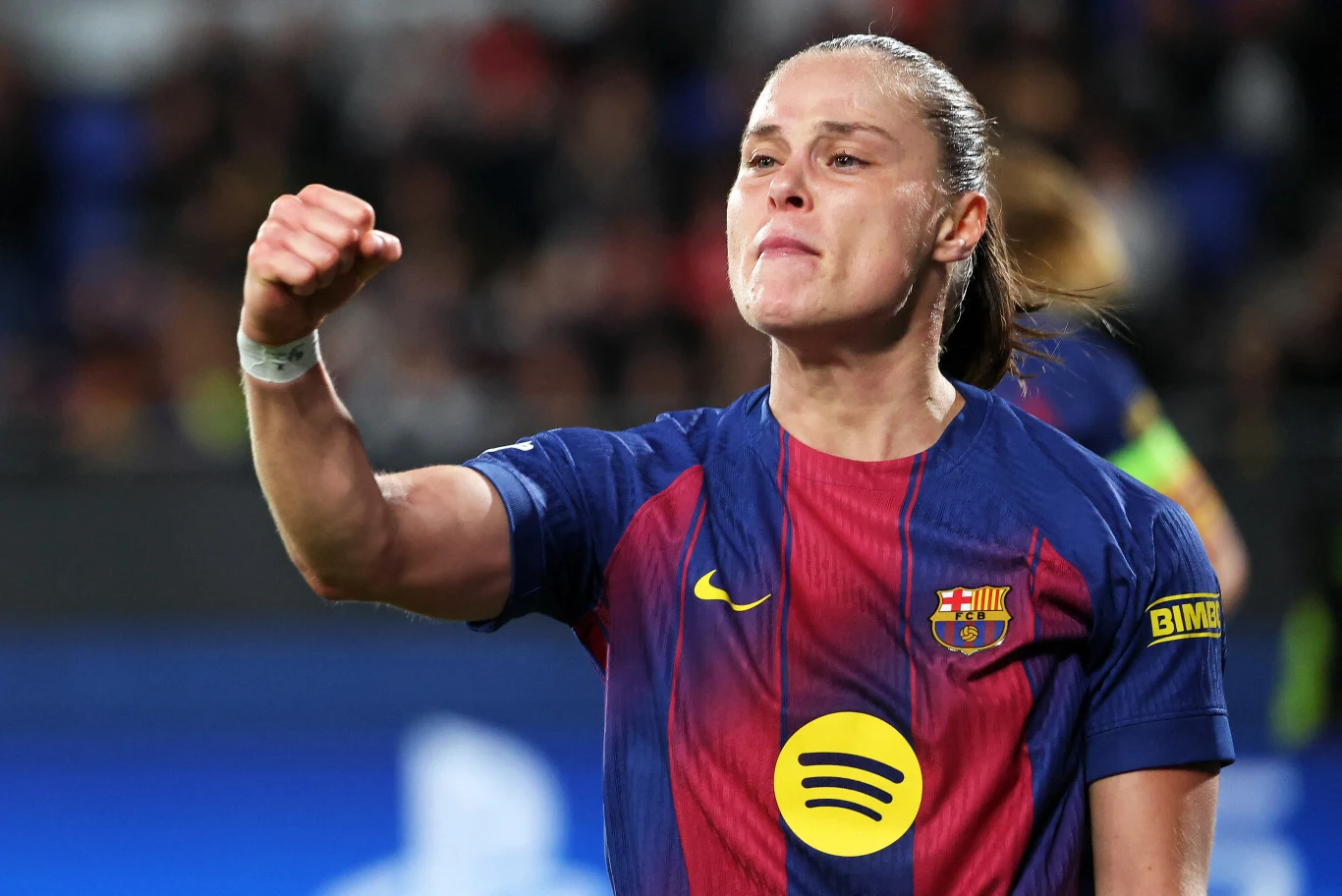 Zawodniczka drużyny FC Barcelona w stroju sportowym z widocznym logotypem klubu oraz sponsora, wykonuje gest triumfu unosząc zaciśniętą pięść, na jej twarzy widać silne emocje i determinację.