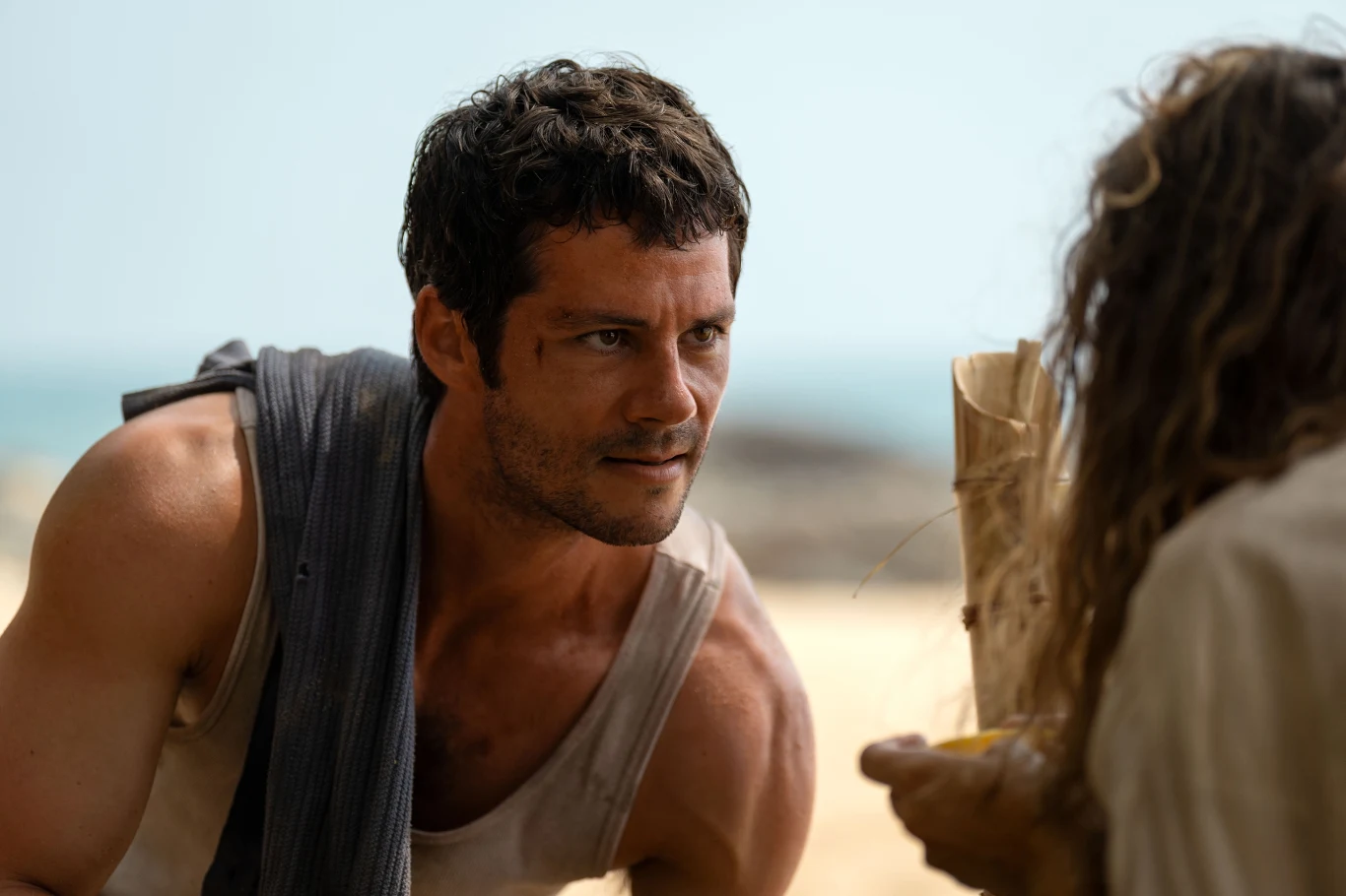 Mężczyzna o umięśnionej sylwetce w jasnej koszulce, pochylony i skupiony, z niewielką raną na twarzy, patrzy na drugą osobę, która trzyma w rękach papierową torbę i żółty przedmiot. W tle widoczna rozmyta plaża i morze.