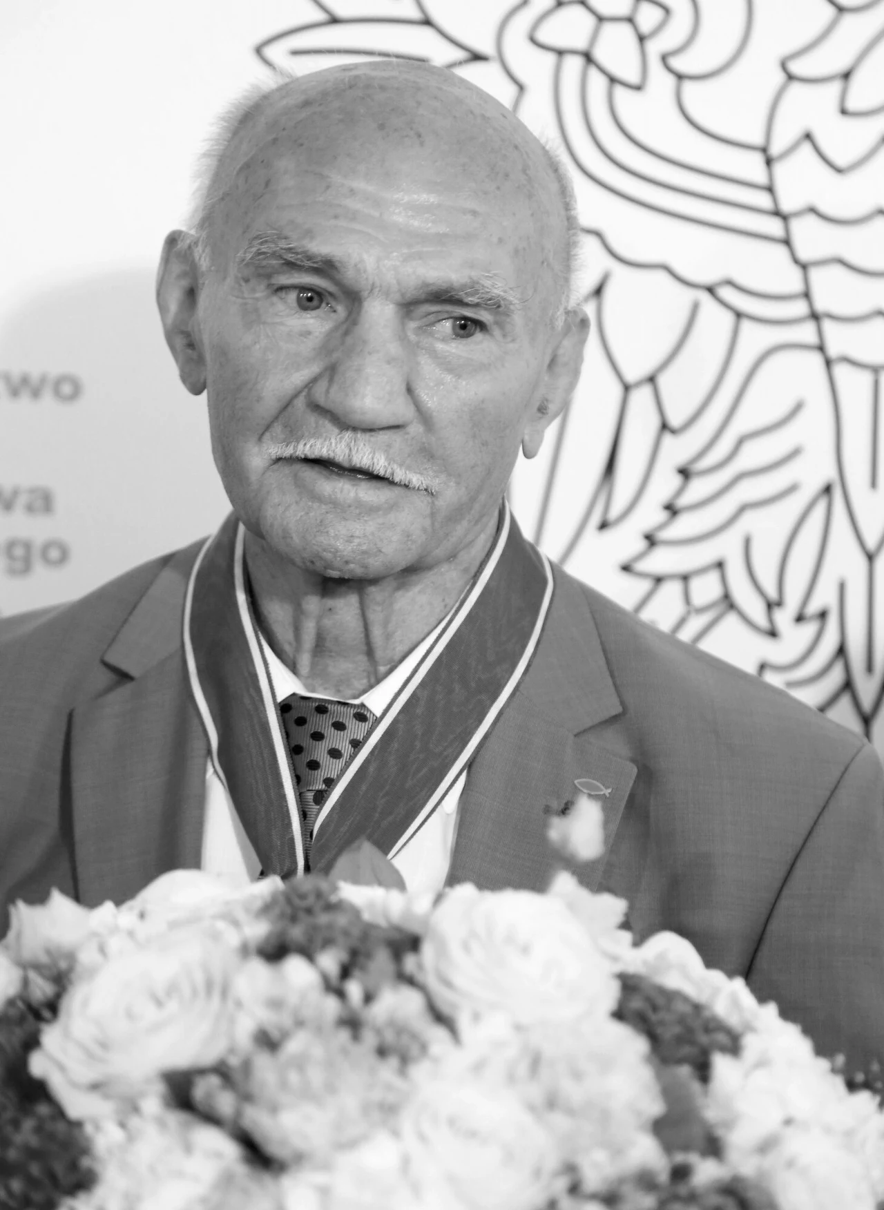 Starszy mężczyzna w garniturze z medalem na szyi, trzyma bukiet kwiatów. W tle widoczny fragment dekoracyjnej grafiki z orłem.
