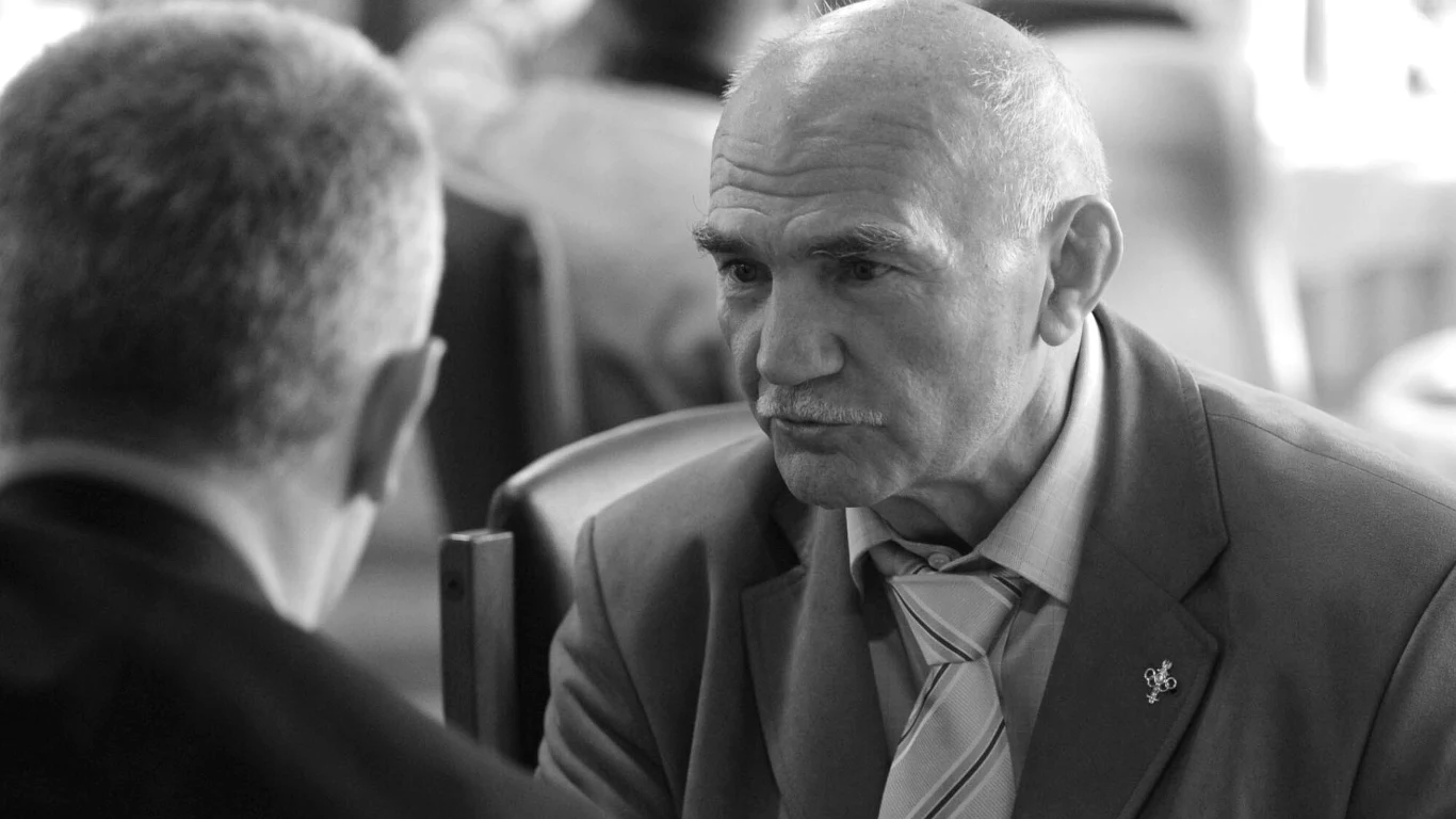 Dwóch starszych mężczyzn rozmawia ze sobą przy stole. Jeden z nich ujęty en face, skupiony, w garniturze i krawacie, z przypinką na klapie.