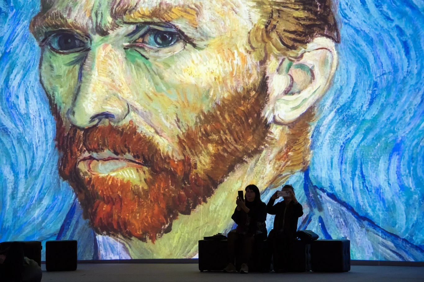 Vincent van Gogh to jeden z największych malarzy w historii Olbrzymie powiększenie słynnego autoportretu Vincenta van Gogha z charakterystycznym ekspresyjnym pociągnięciem pędzla i intensywnymi barwami, na tle którego dwie osoby siedzą na ławkach w cieniu przy podświetlonym ekranie.