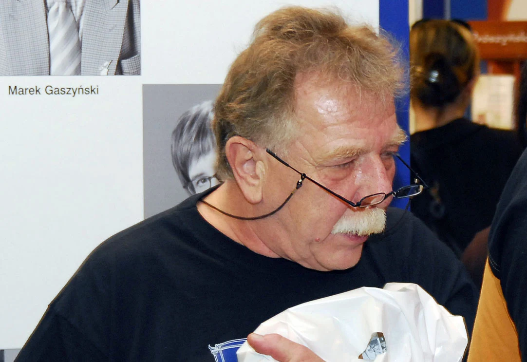 Piotr Kaczkowski od lat unika publikowania swoich fotografii w mediach (to zdjęcie z 2006 r.) Starszy mężczyzna z wąsem i okularami opartymi na nosie trzyma w ręku biały pakunek. W tle rozpoznawalne wydrukowane fotografie innych osób oraz fragmenty napisu.