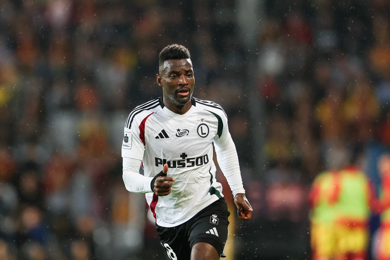 Piłkarz w biało-czarnej koszulce drużyny Legii Warszawa biegnie po boisku w trakcie meczu, w tle widać zamazaną publiczność oraz innych zawodników.