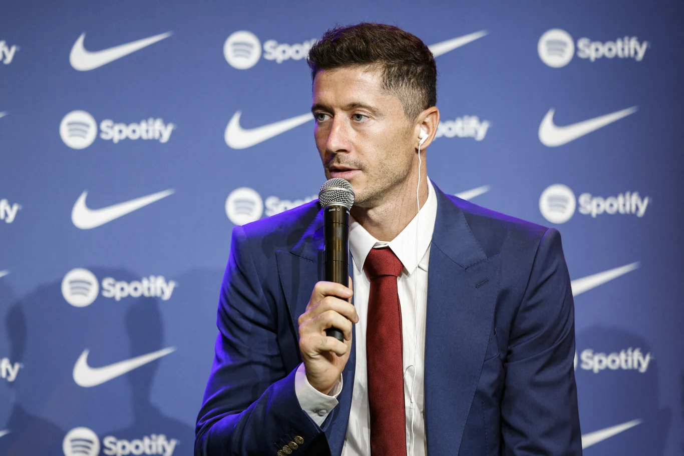 Robert Lewandowski Mężczyzna w niebieskim garniturze i czerwonym krawacie siedzi na tle z logo Spotify i Nike, trzyma mikrofon, przygotowany do wypowiedzi lub wywiadu.