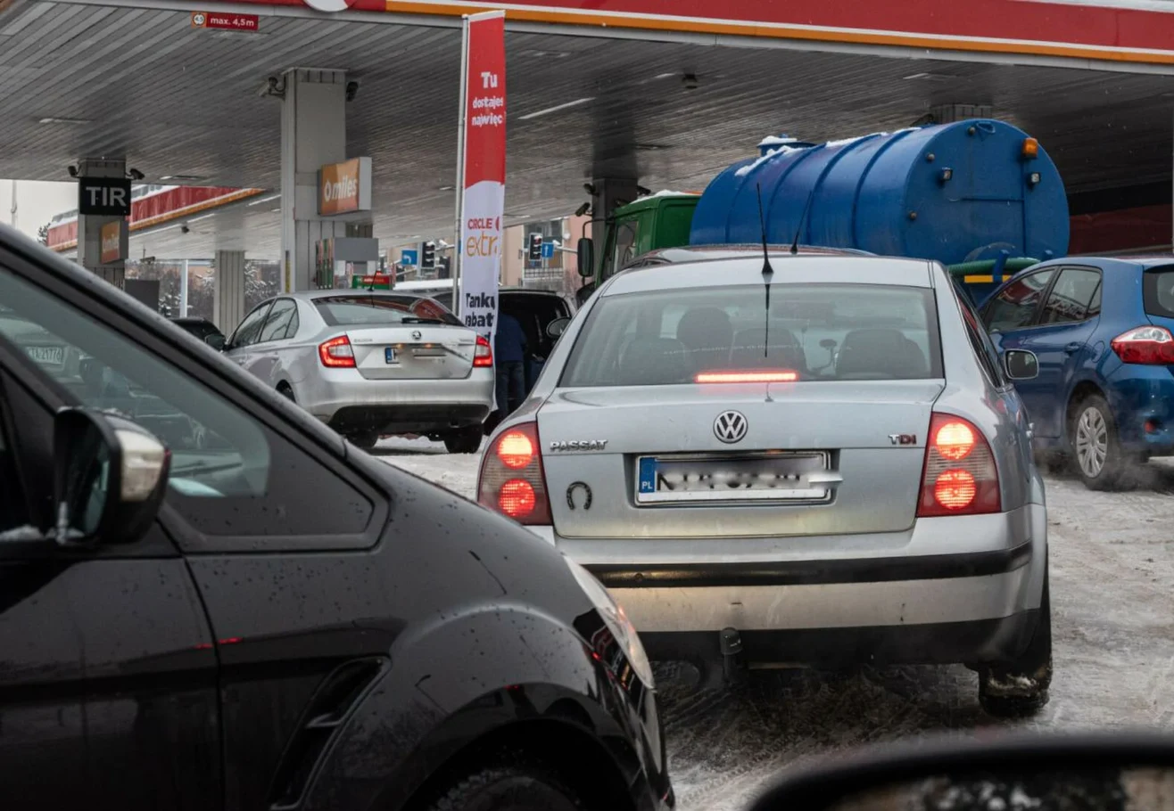 Kolejka samochodów przy stacji benzynowej w zimowej aurze, widoczny Volkswagen z tyłu, niebieska cysterna oraz zadaszenie stacji paliw.