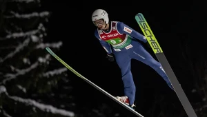 Zawodnik w stroju skoczka narciarskiego unosi się w powietrzu podczas nocnego konkursu na tle ciemnych drzew, widoczny numer startowy i sprzęt sportowy.