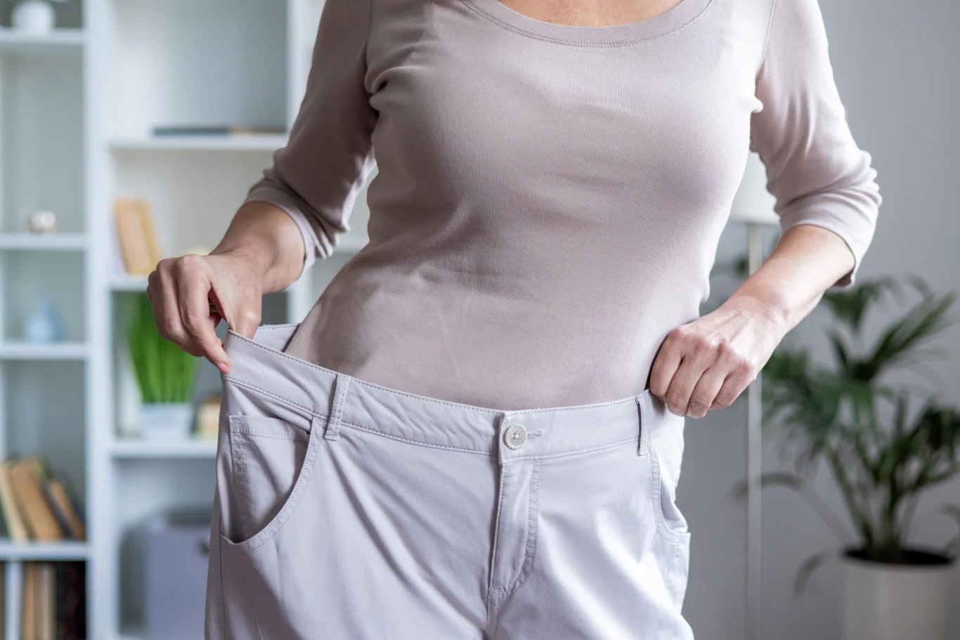 Nagła utrata masy ciała, nawracające bóle brzucha i zmiana rytmu wypróżnień to objawy nowotworu przewodu pokarmowego Osoba trzyma wyraźnie za duże spodnie za pasek, ubrana w jasną bluzkę z długim rękawem, stoi w jasnym, domowym wnętrzu z rośliną oraz półkami w tle.