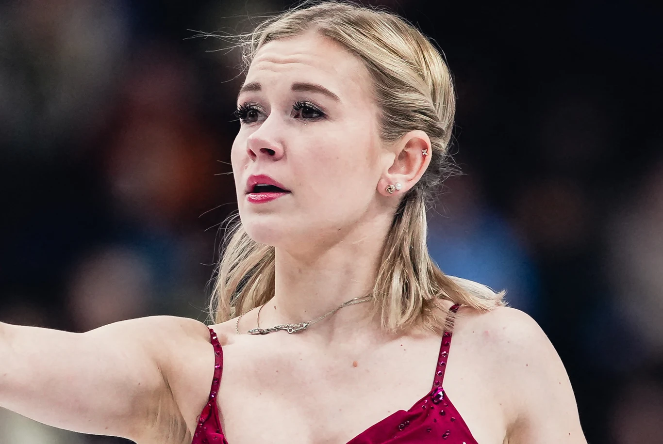 Młoda kobieta w stroju sportowym na tle rozmytej publiczności, z poważnym wyrazem twarzy oraz uniesioną ręką, prawdopodobnie podczas występu na lodzie.