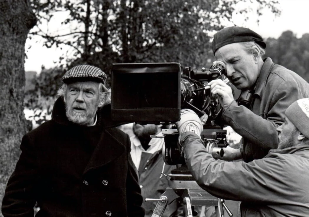 Dwóch mężczyzn pracuje przy kamerze filmowej na zewnątrz, jeden spogląda przez wizjer kamery, drugi stoi obok i patrzy w bok, scena utrzymana w czerni i bieli, w tle widoczne drzewa.